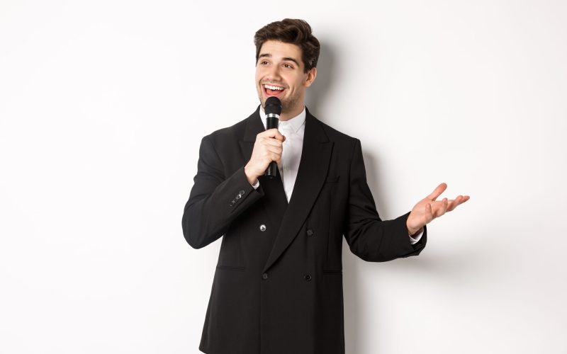 Portrait of handsome man in black suit singing a song, holding microphone and giving speech, standing against white background.